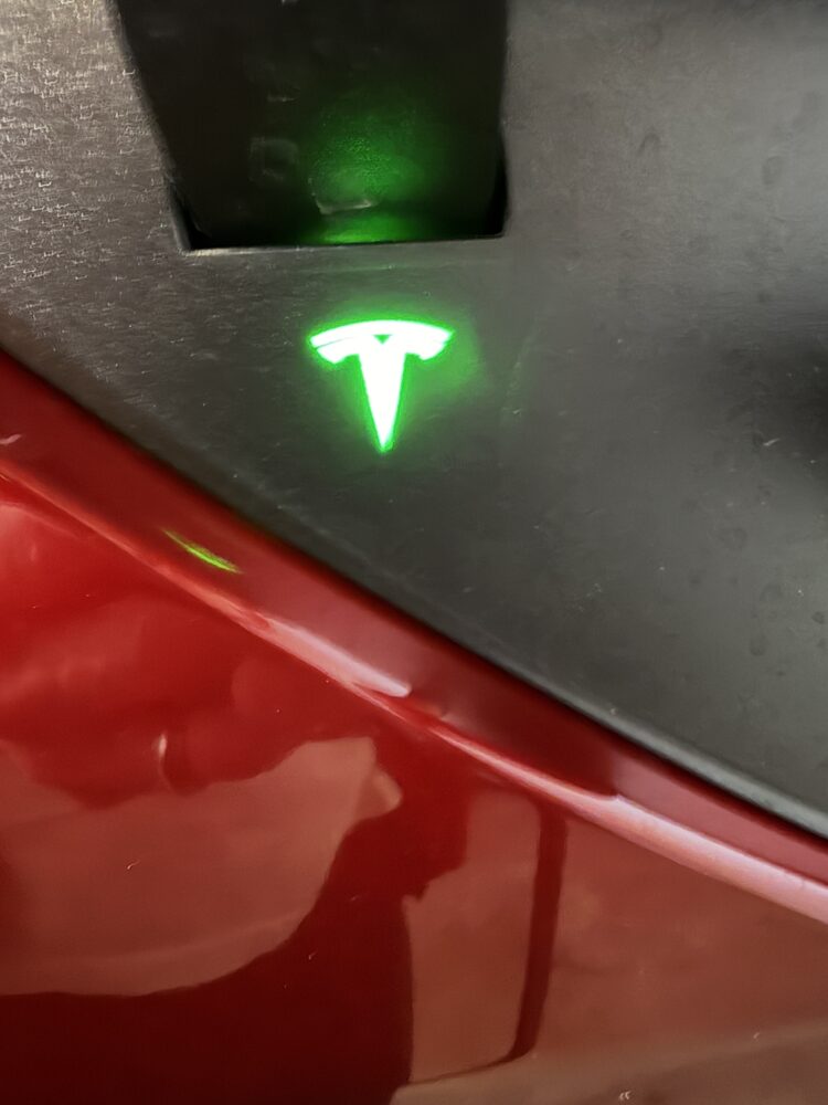 Tesla charge port with the T logo glowing green while the car charges.
