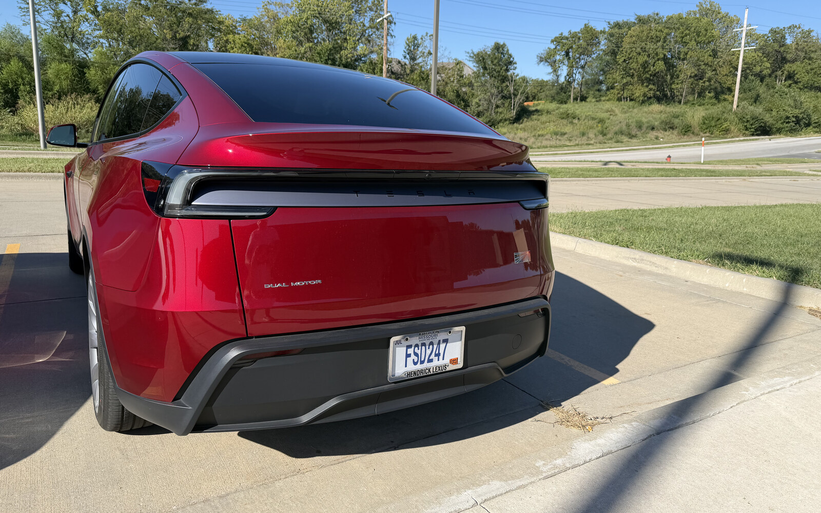 Rear three-quarter view of a Midnight Cherry Red Tesla Model Y parked on concrete under a clear sky, wearing the Missouri plate FSD247, with trees and power lines behind it.