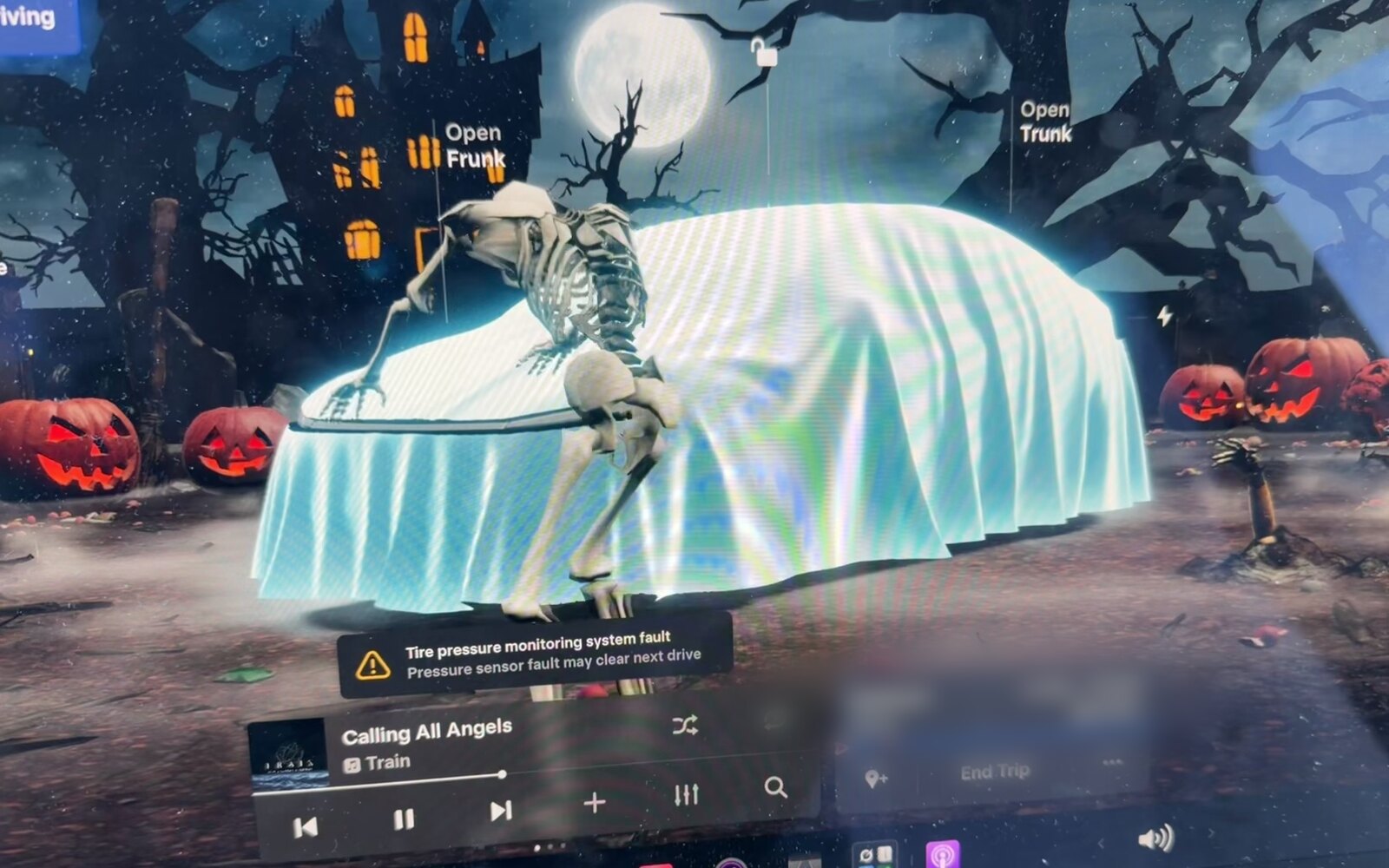 Tesla touchscreen in Halloween mode: the Model Y drawn as a ghost under a white sheet in front of a haunted house, a full moon and jack-o'-lanterns, with a skeleton walking around the car.