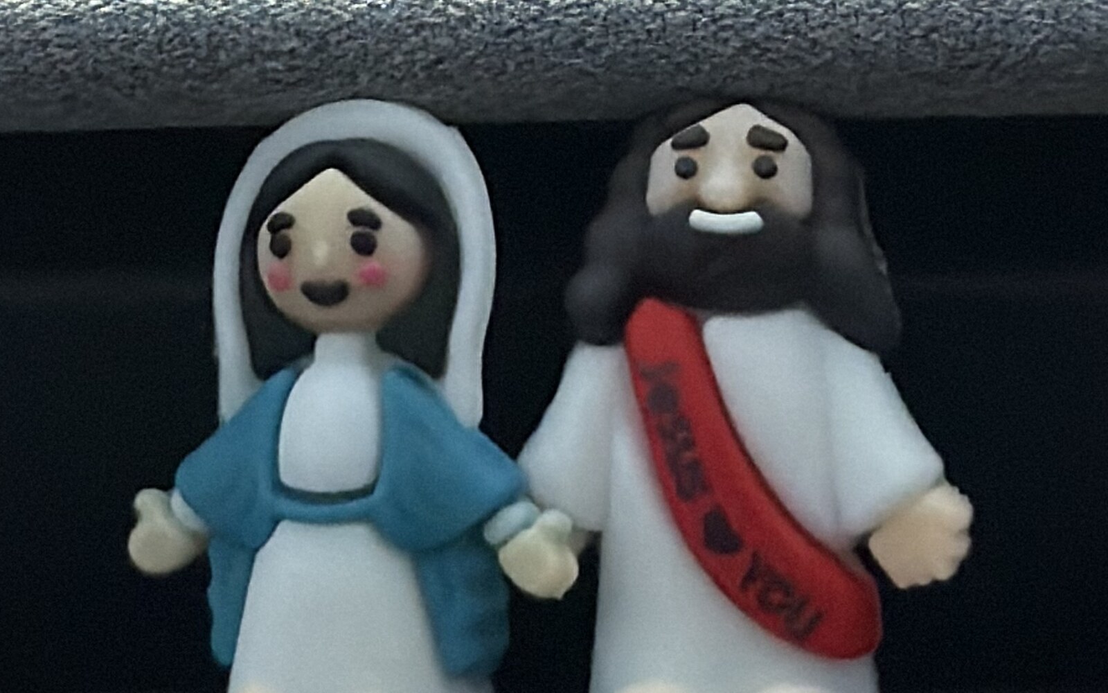 Two small figurines, Mary and Jesus, standing side by side beneath the dashboard lip of a Tesla.