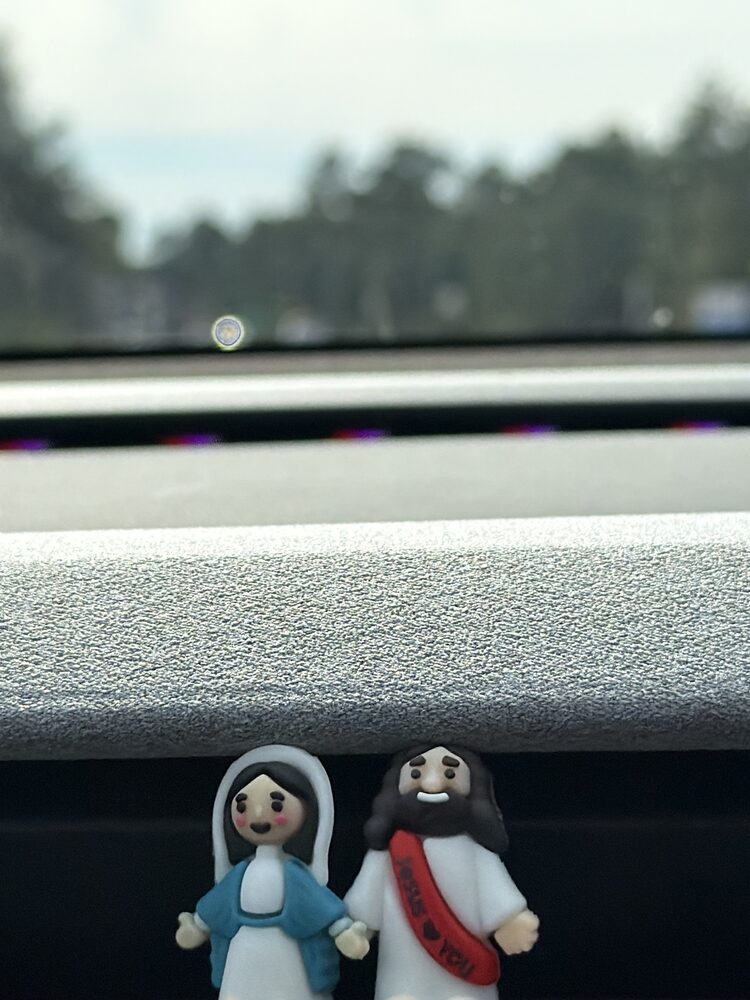 Two small figurines of Jesus and Mary standing on the dashboard of a Tesla with the road blurred beyond the windshield.