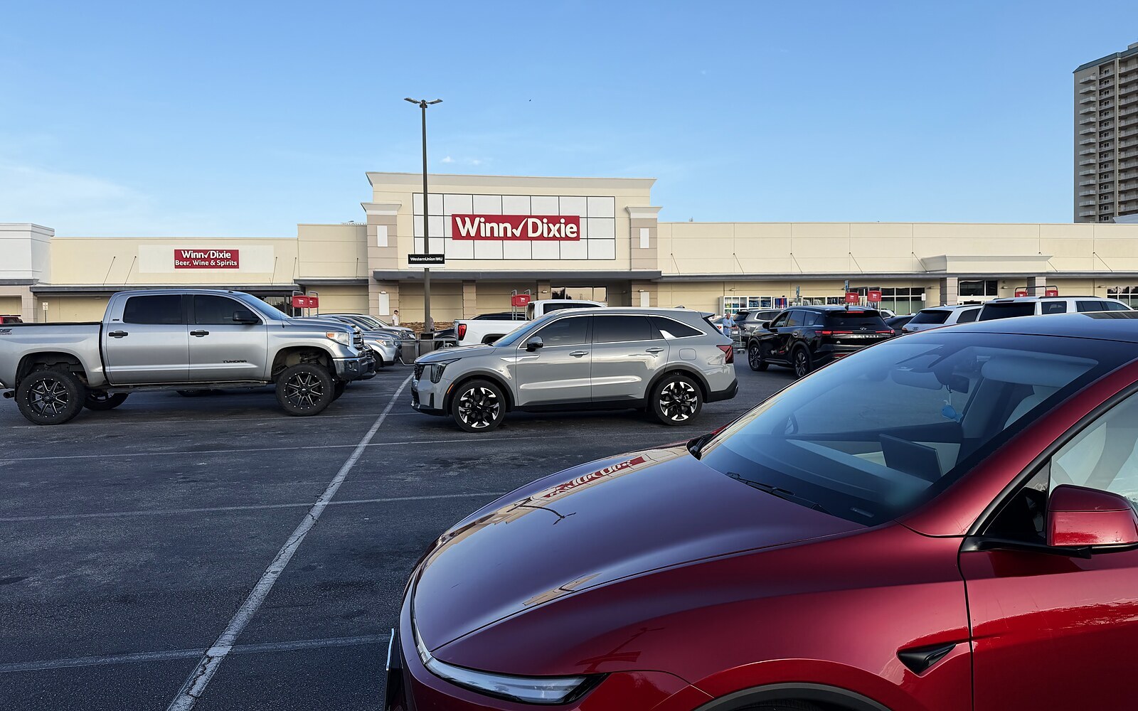 Red Tesla Model Y parked alone in an empty row of a Winn-Dixie lot on the Florida coast, with the store and other cars far away.
